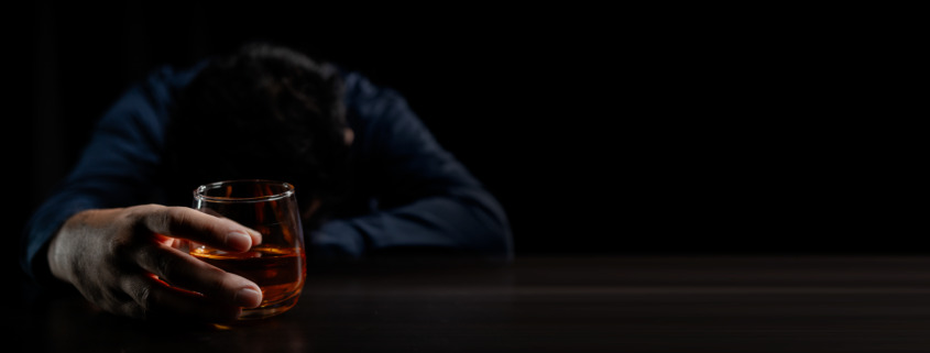 A man with his head down on a table holding a glass of alcohol wondering what are the best detox programs for alcohol.